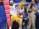 LSU football player wearing #14 in white jersey and yellow pants makes a sideline catch, with photographers and fans watching in the background.
