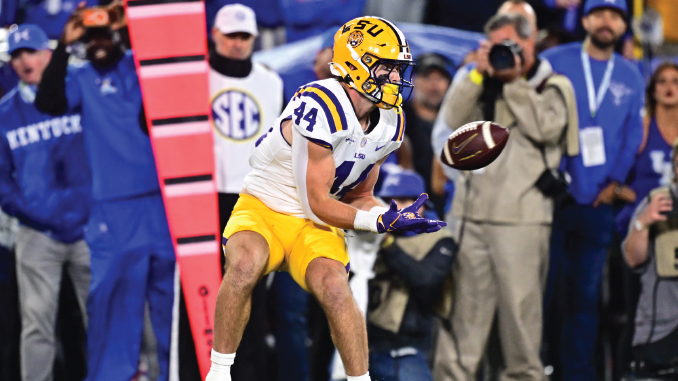 LSU football player wearing #14 in white jersey and yellow pants makes a sideline catch, with photographers and fans watching in the background.