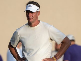 Coach wearing an LSU visor and white athletic shirt stands outdoors with hands on hips.
