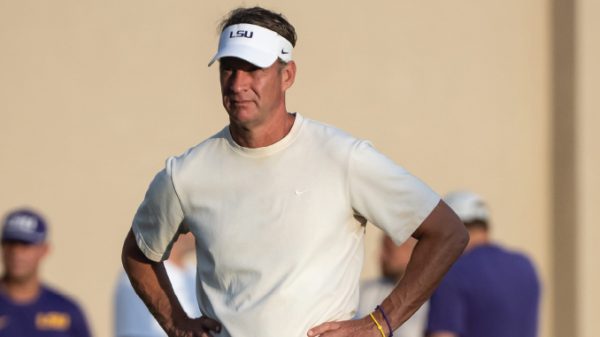Coach wearing an LSU visor and white athletic shirt stands outdoors with hands on hips.