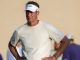 Coach wearing an LSU visor and white athletic shirt stands outdoors with hands on hips.