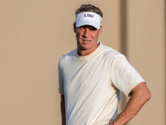 Man on a golf course wearing a white short-sleeve shirt and an LSU-branded white visor, hands on hips.