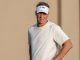 Man on a golf course wearing a white short-sleeve shirt and an LSU-branded white visor, hands on hips.