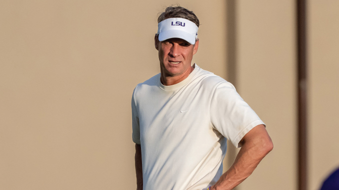 Man on a golf course wearing a white short-sleeve shirt and an LSU-branded white visor, hands on hips.
