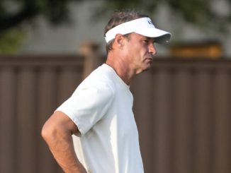 Man in a white polo and white visor stands outdoors, facing to the right with a thoughtful expression.