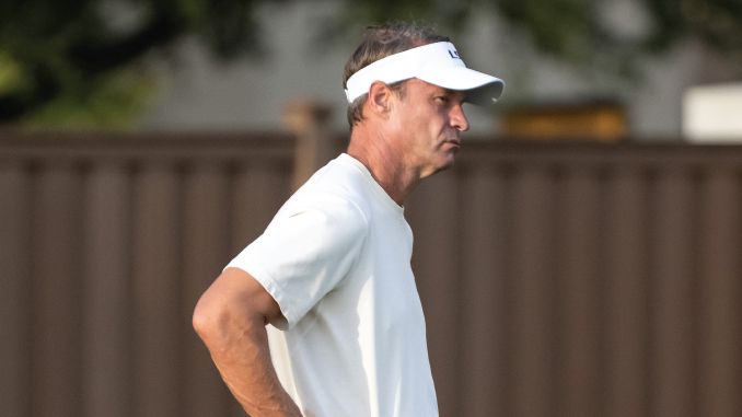 Man in a white polo and white visor stands outdoors, facing to the right with a thoughtful expression.