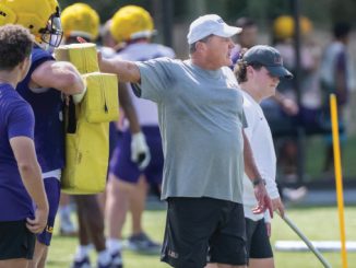 Coach directing a football drill on a sunny field, pointing as players in purple and yellow gear practice nearby with yellow blocking pads nearby