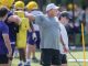 Coach directing a football drill on a sunny field, pointing as players in purple and yellow gear practice nearby with yellow blocking pads nearby