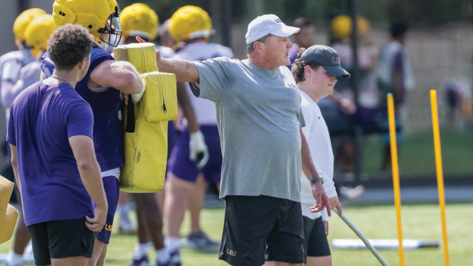 Coach directing a football drill on a sunny field, pointing as players in purple and yellow gear practice nearby with yellow blocking pads nearby