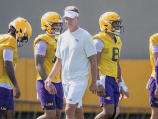 LSU football coach in white attire walks with players wearing yellow helmets during practice on a sunny day.