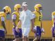 LSU football coach in white attire walks with players wearing yellow helmets during practice on a sunny day.