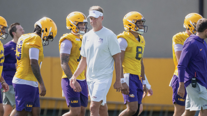 LSU football coach in white attire walks with players wearing yellow helmets during practice on a sunny day.