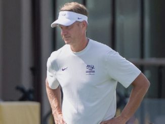 Man in a white athletic shirt and visor stands outdoors with hands on hips, looking to the side near a training facility.