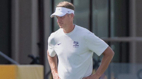 Man in a white athletic shirt and visor stands outdoors with hands on hips, looking to the side near a training facility.