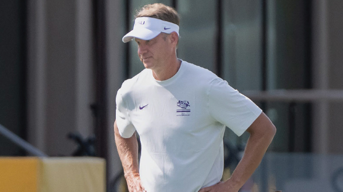 Man in a white athletic shirt and visor stands outdoors with hands on hips, looking to the side near a training facility.