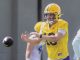 Football player in a yellow helmet and jersey stretching to catch a pass during a practice session, with teammates in the background.