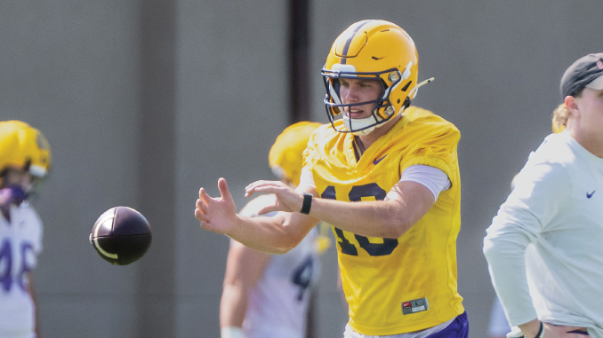 Football player in a yellow helmet and jersey stretching to catch a pass during a practice session, with teammates in the background.