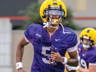 A college football player in a purple jersey with the number 5 and a yellow helmet runs on the field, with another purple-jersey teammate in the background.