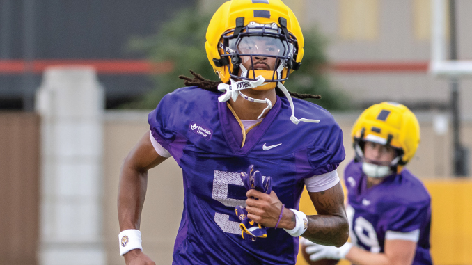 A college football player in a purple jersey with the number 5 and a yellow helmet runs on the field, with another purple-jersey teammate in the background.