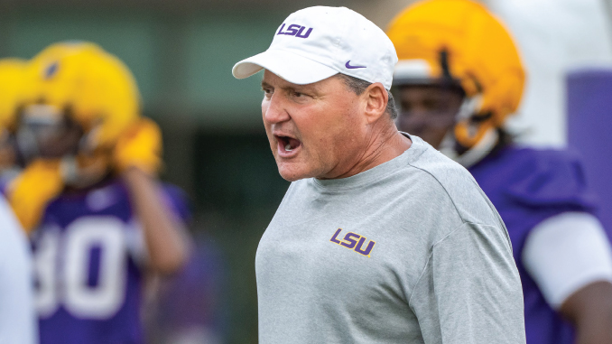 LSUFball_Practice080626_EdOrgeron | TigerRag.com A football coach in an LSU gray shirt and white cap shouting during practice, with players in yellow helmets in the background.