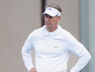 Man wearing a white LSU visor and hoodie stands with hands on hips, looking focused outside.