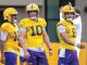 Three LSU football players in yellow practice uniforms stand on a field, center #10 flanked by teammates #15 and #8.