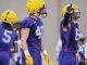 Football players in purple jerseys and yellow helmets during practice; center player wearing #41 stands with glove-clad hands, others in motion behind him.