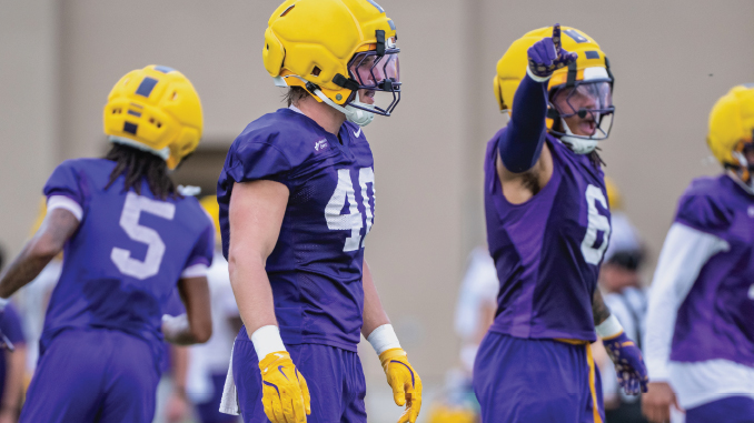 Football players in purple jerseys and yellow helmets during practice; center player wearing #41 stands with glove-clad hands, others in motion behind him.