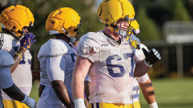 Football players in yellow helmets and white uniforms on a sunny practice field; foreground player wearing number 67.