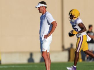 Coach in a white outfit and visor stands with hands on hips watching football practice, LSU player in purple and gold in the background.