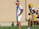 Coach in a white outfit and visor stands with hands on hips watching football practice, LSU player in purple and gold in the background.
