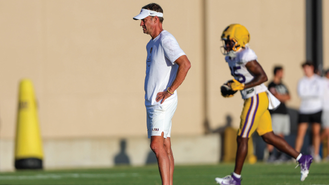 Coach in a white outfit and visor stands with hands on hips watching football practice, LSU player in purple and gold in the background.
