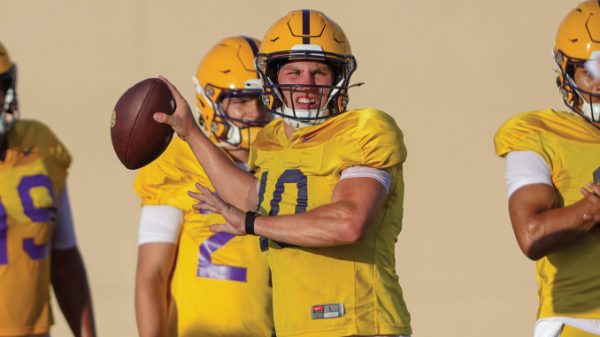 Quarterback in a yellow jersey throws a pass during drills, teammates in the background wearing matching uniforms.