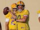 Quarterback in a yellow jersey throws a pass during drills, teammates in the background wearing matching uniforms.