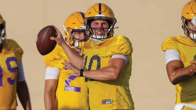 Quarterback in a yellow jersey throws a pass during drills, teammates in the background wearing matching uniforms.