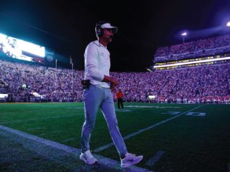 Football coach walking across the field at night beneath stadium lights, headset on, with a packed crowd in the stands.