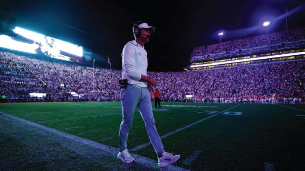 Football coach walking across the field at night beneath stadium lights, headset on, with a packed crowd in the stands.