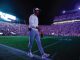 Football coach walking across the field at night beneath stadium lights, headset on, with a packed crowd in the stands.