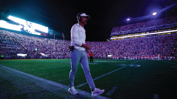 Football coach walking across the field at night beneath stadium lights, headset on, with a packed crowd in the stands.