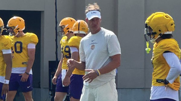 LSU football coach in a light gray shirt and visor watches players in yellow helmets and jerseys during outdoor practice, with drills near a training facility; visible jersey numbers include 80 and 59.