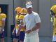 LSU football coach in a light gray shirt and visor watches players in yellow helmets and jerseys during outdoor practice, with drills near a training facility; visible jersey numbers include 80 and 59.