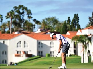 Golfer in a white cap and polo lines up a putt on a green with white, red-roofed buildings and palm trees in the background.