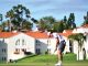 Golfer in a white cap and polo lines up a putt on a green with white, red-roofed buildings and palm trees in the background.