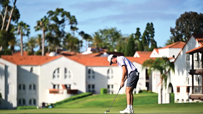 Golfer in a white cap and polo lines up a putt on a green with white, red-roofed buildings and palm trees in the background.
