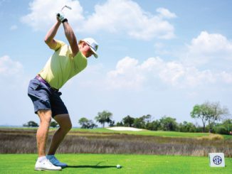 Golfer in a lime shirt and white cap mid-swing on a sunny golf course, tee ball at his feet.
