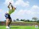 Golfer in a lime shirt and white cap mid-swing on a sunny golf course, tee ball at his feet.