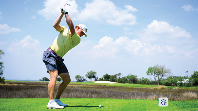 Golfer in a lime shirt and white cap mid-swing on a sunny golf course, tee ball at his feet.
