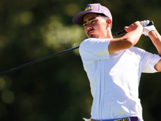 Male golfer in a white polo swings a club on a sunny golf course, wearing a purple LSU cap.