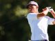 Male golfer in a white polo swings a club on a sunny golf course, wearing a purple LSU cap.