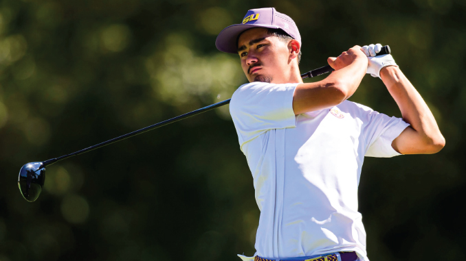 Male golfer in a white polo swings a club on a sunny golf course, wearing a purple LSU cap.